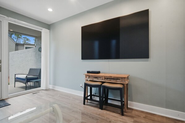A room with a large flat-screen TV mounted on the wall, a wooden table with two stools, and a glass door leading to an outdoor area with a chair. - 1259 Harbour Town Club at The Sea Pines Resort (Hilton Head Island)