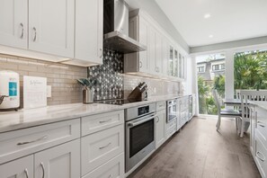 A modern kitchen with white cabinets, a stainless steel range hood, a tiled backsplash, and a view of the outdoors.