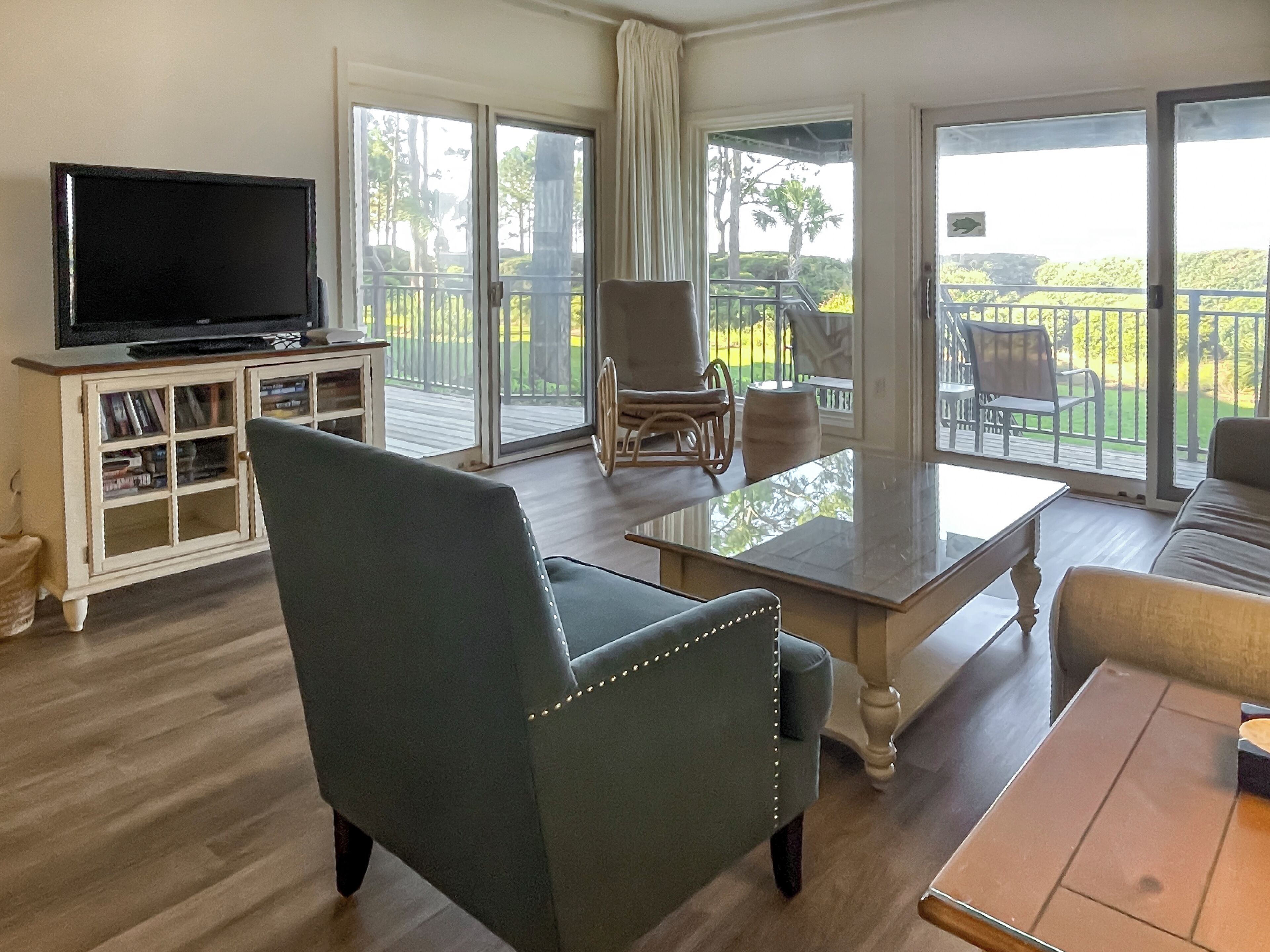 Un salón amplio con televisor, estantería, sillón verde, mesa de centro y balcón con mobiliario exterior.
