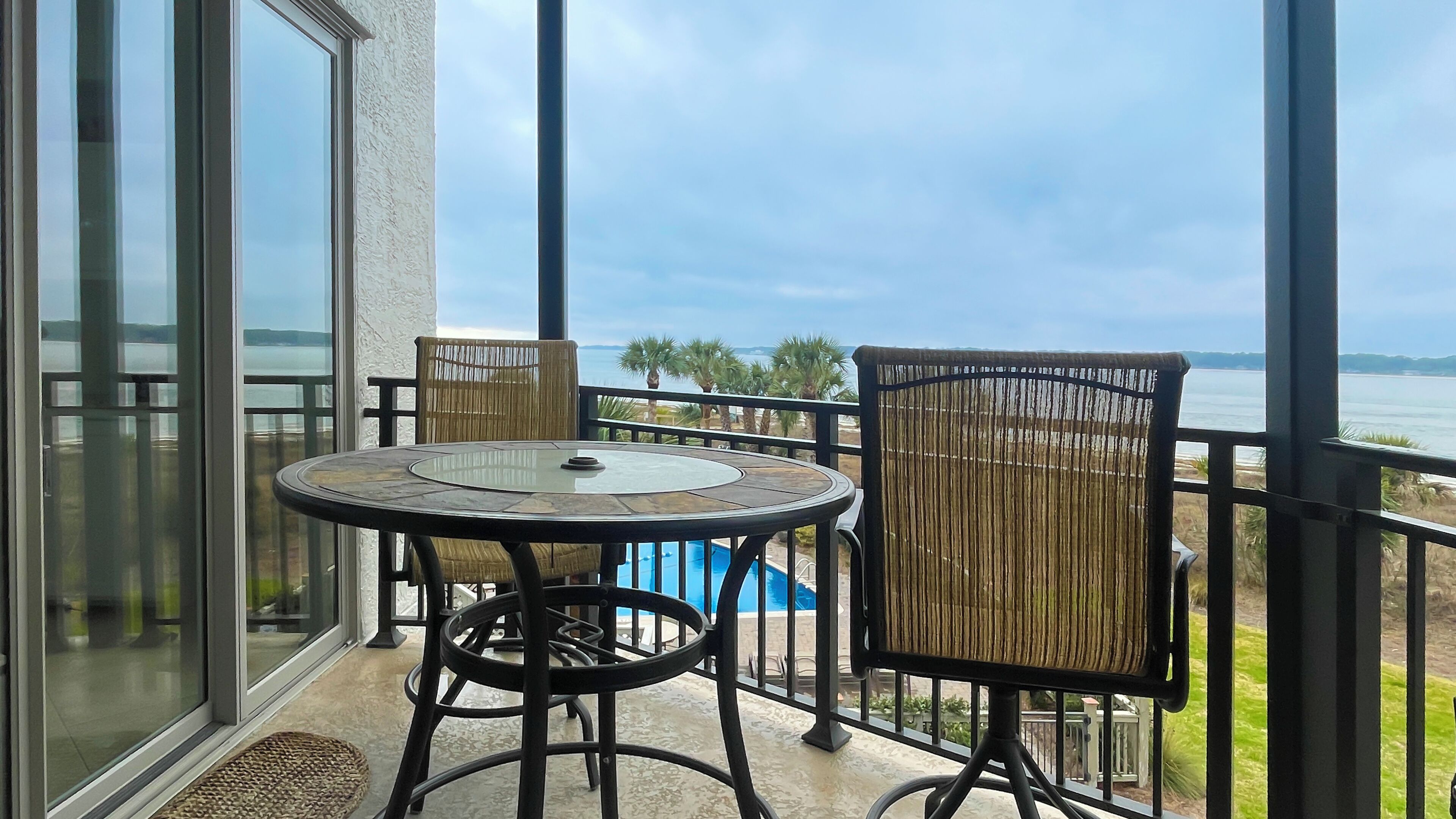 A balcony with a glass door, a round table with chairs, and a view of the sea.