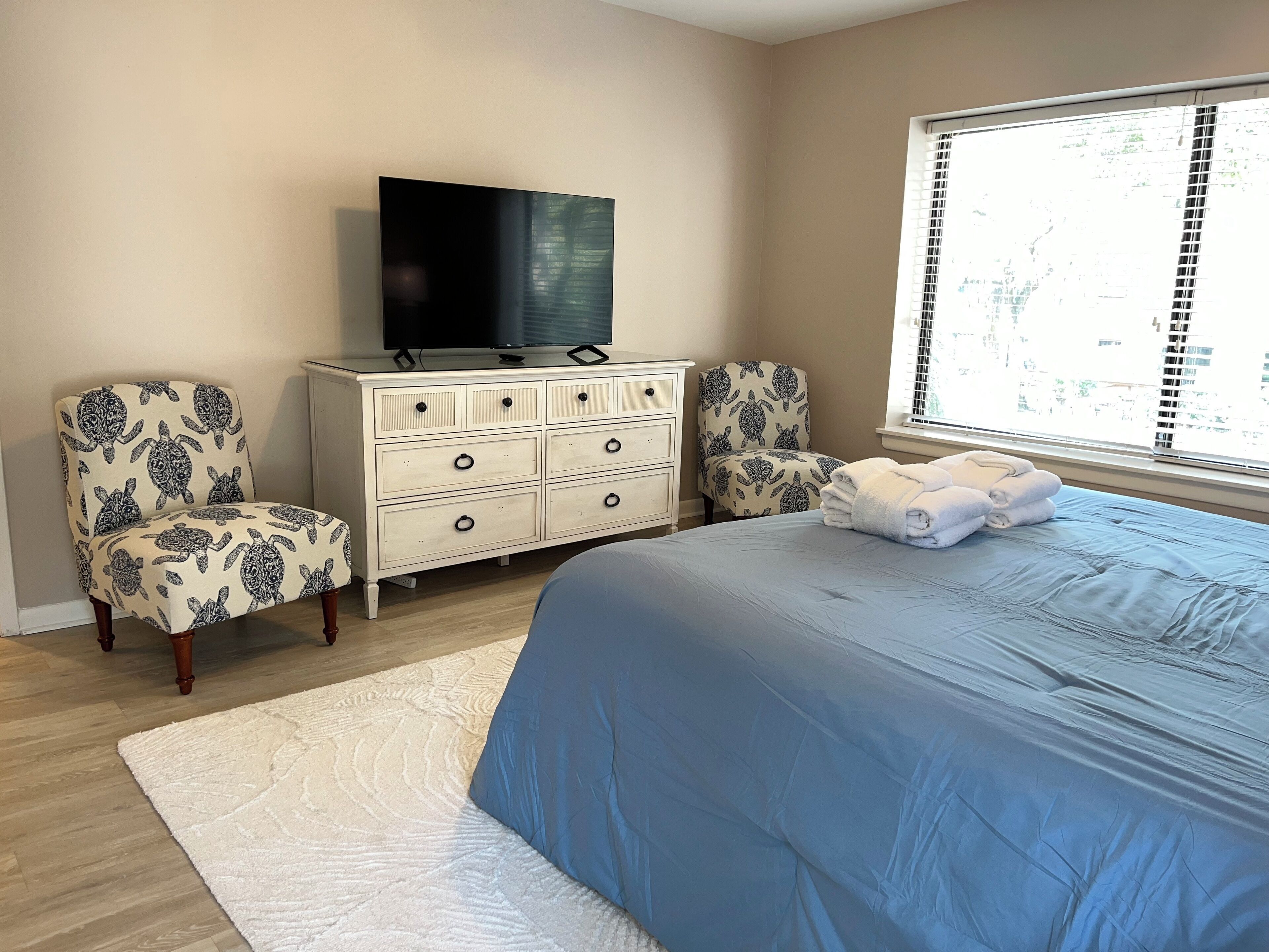 A bedroom with a bed, a dresser, two patterned chairs, a television, and a window with blinds.