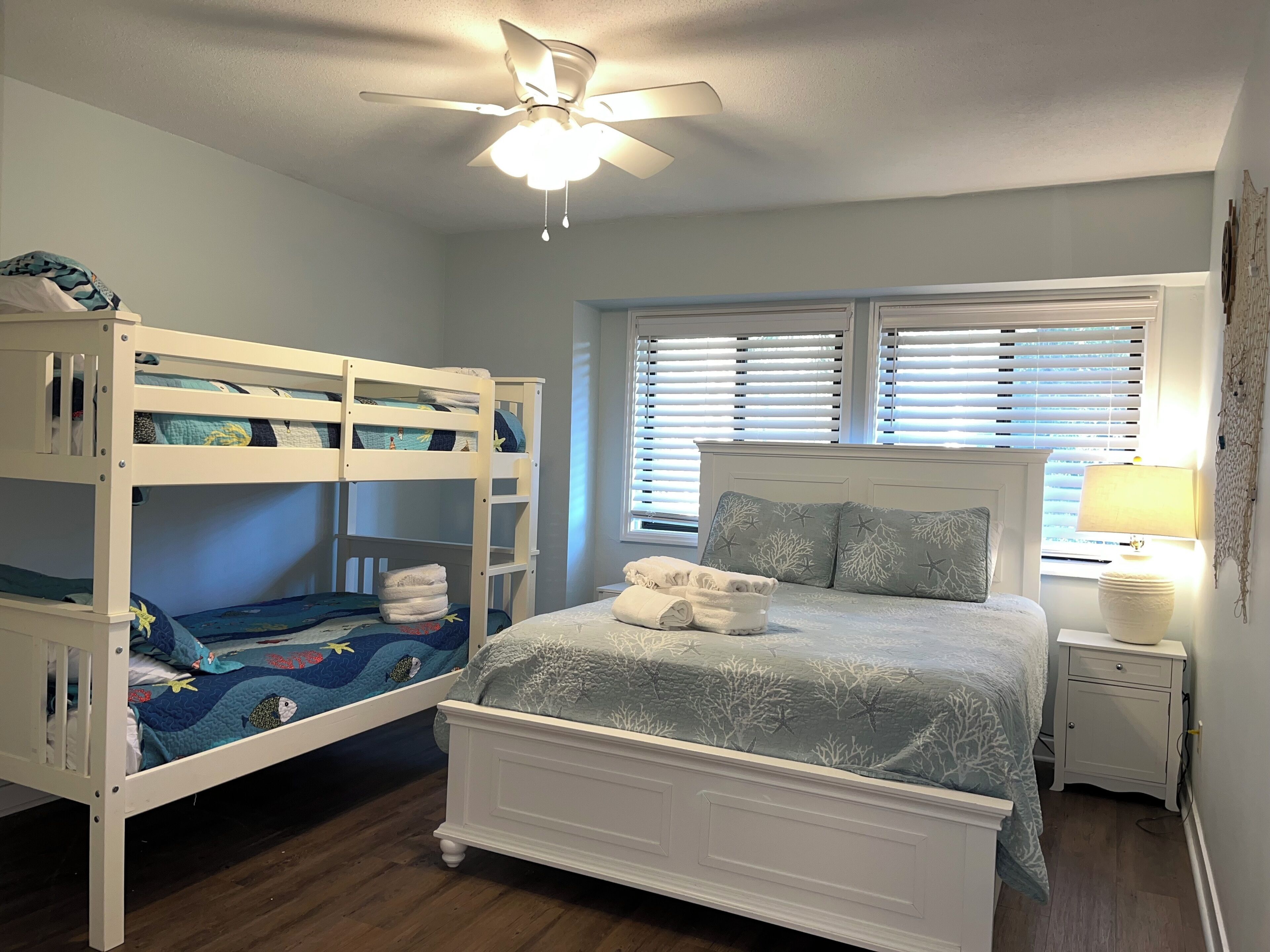 A bedroom with a bunk bed, a single bed, a ceiling fan, a window with blinds, a bedside table with a lamp, and a nightstand.