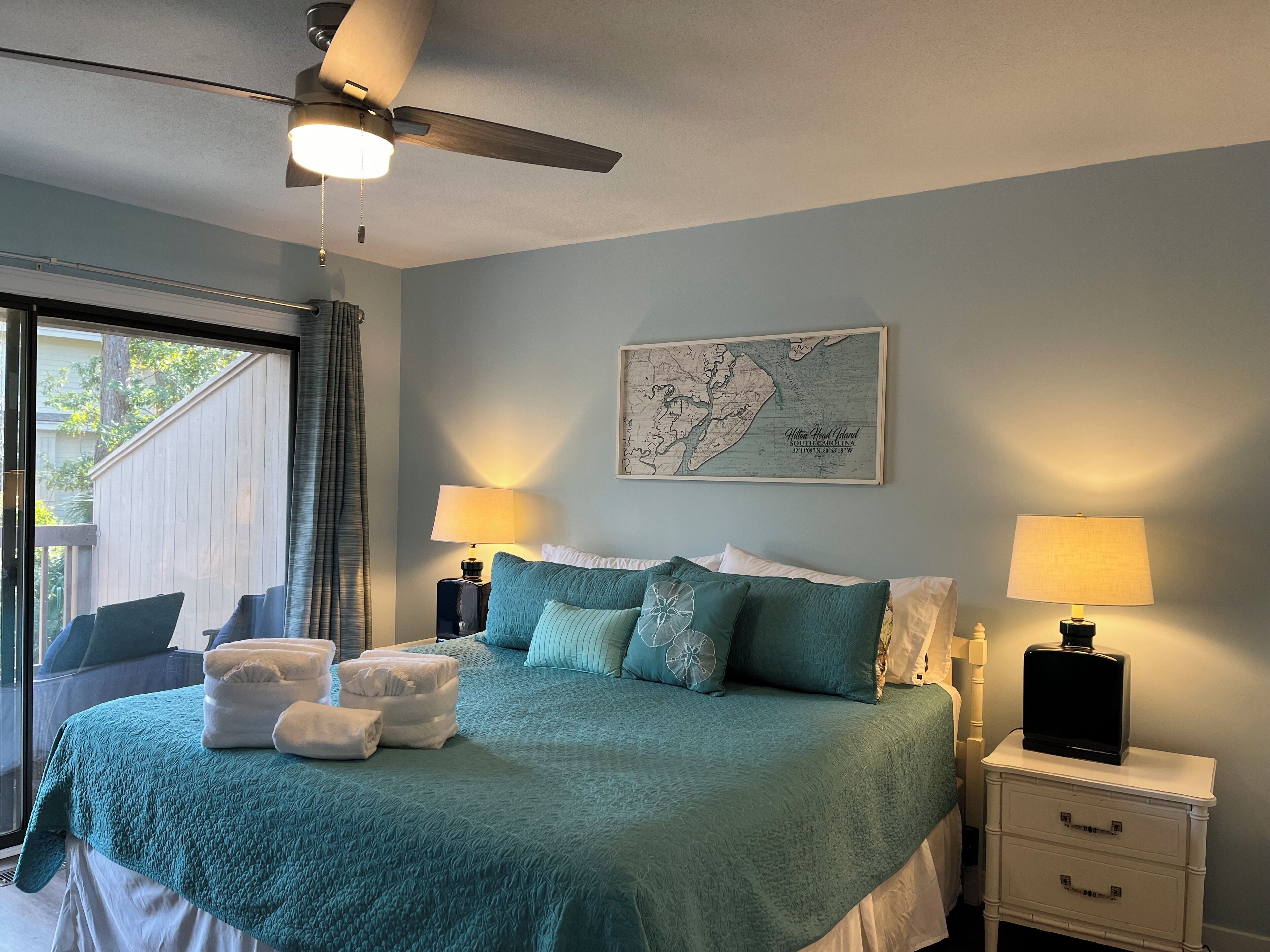Une chambre à coucher avec un lit, deux tables de chevet, un ventilateur de plafond, une carte accrochée au mur et une vue sur l’extérieur.