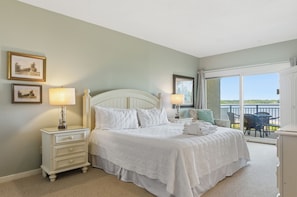 Une chambre à coucher avec un lit, deux tables de chevet avec des lampes, une commode, un balcon avec une table et des chaises, et des tableaux encadrés accrochés au mur.
