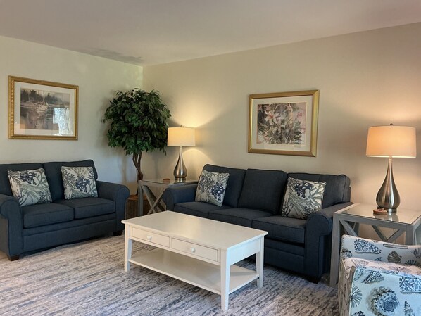 A living room with two dark sofas, a white coffee table, and two lamps. There are framed pictures on the walls and a potted plant. - 2208 Heritage Villas at The Sea Pines Resort (Hilton Head Island)