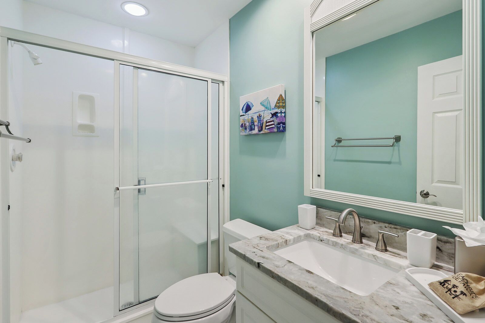 A modern bathroom with a glass shower enclosure, a white toilet, a marble sink with a large mirror, and a teal accent wall.