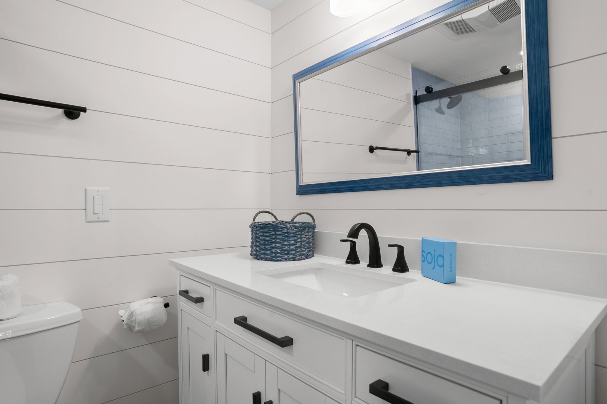 A modern bathroom with white tiled walls, a white sink with black faucets, a blue-framed mirror, and a blue basket on the countertop.