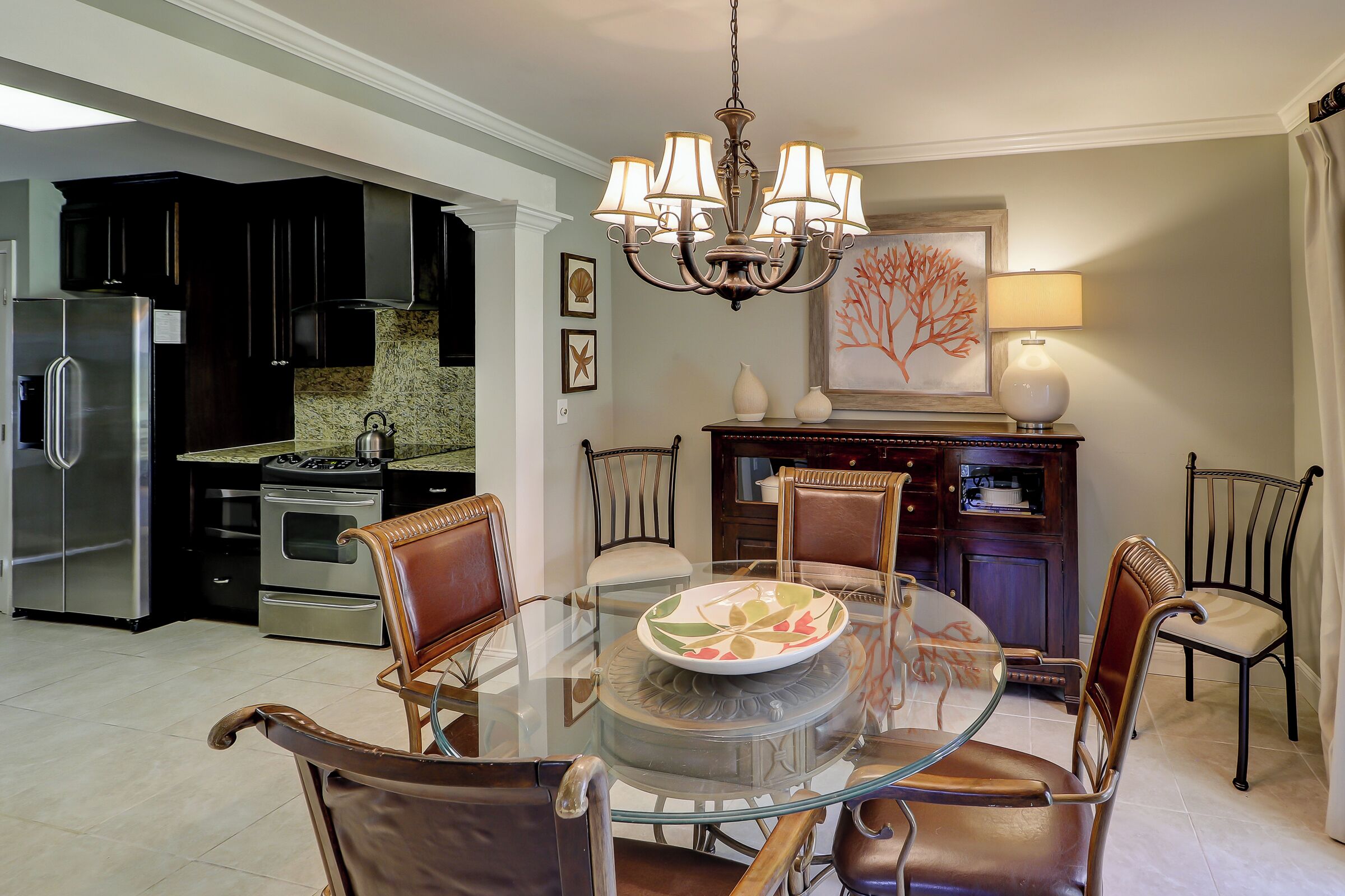 A modern kitchen with stainless steel appliances, a dining area with a glass table and leather chairs, and a chandelier.