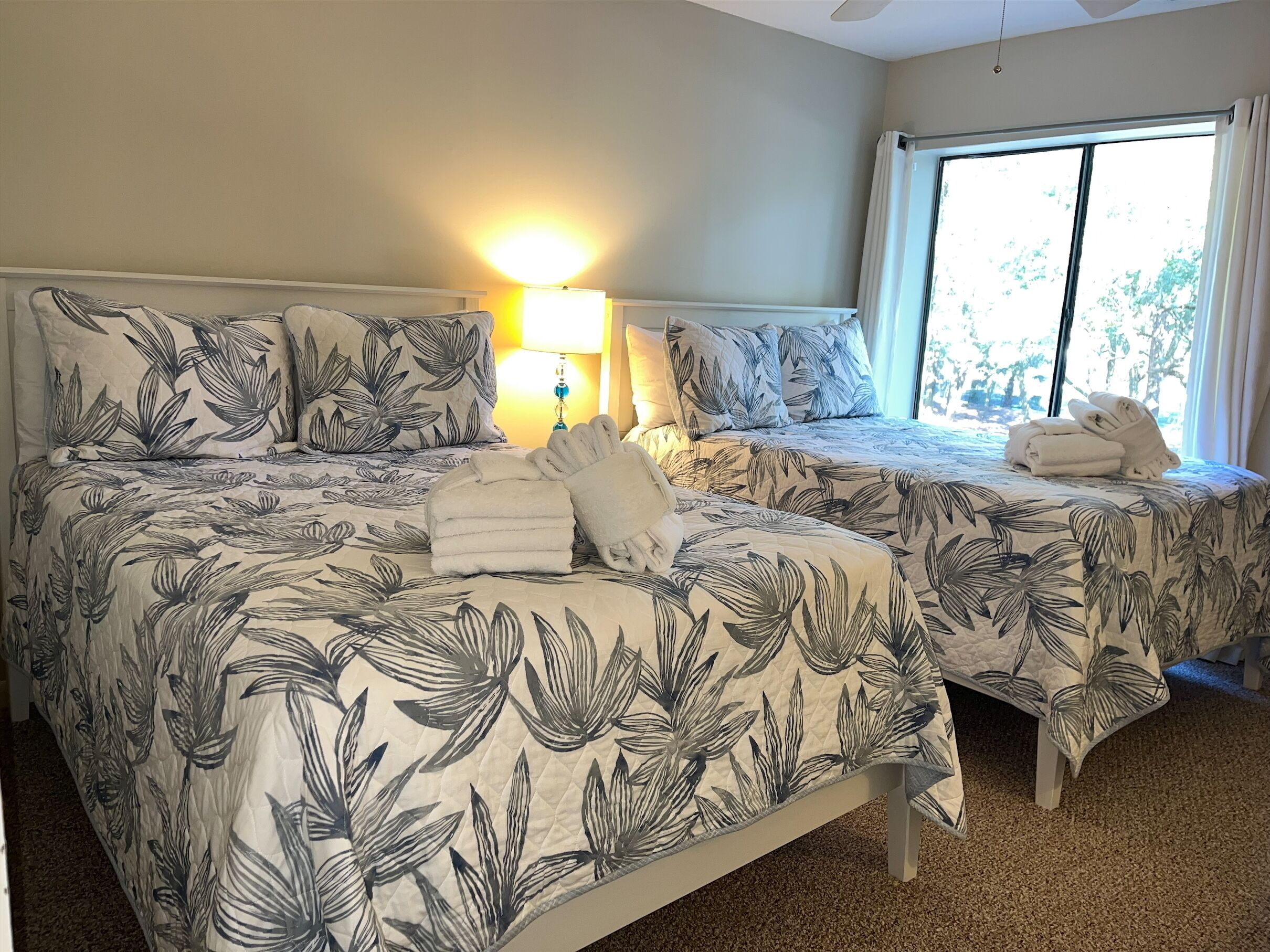 A hotel room with two beds, each with a floral-patterned bedspread, a bedside lamp, and a window with curtains.