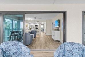 A modern living room with a large sliding glass door leading to a balcony, a kitchen in the background, and a dining area. - Blue Gulf 102 | Gulf-Front 3BR Sanibel Condo with Stunning Views! (Sanibel)