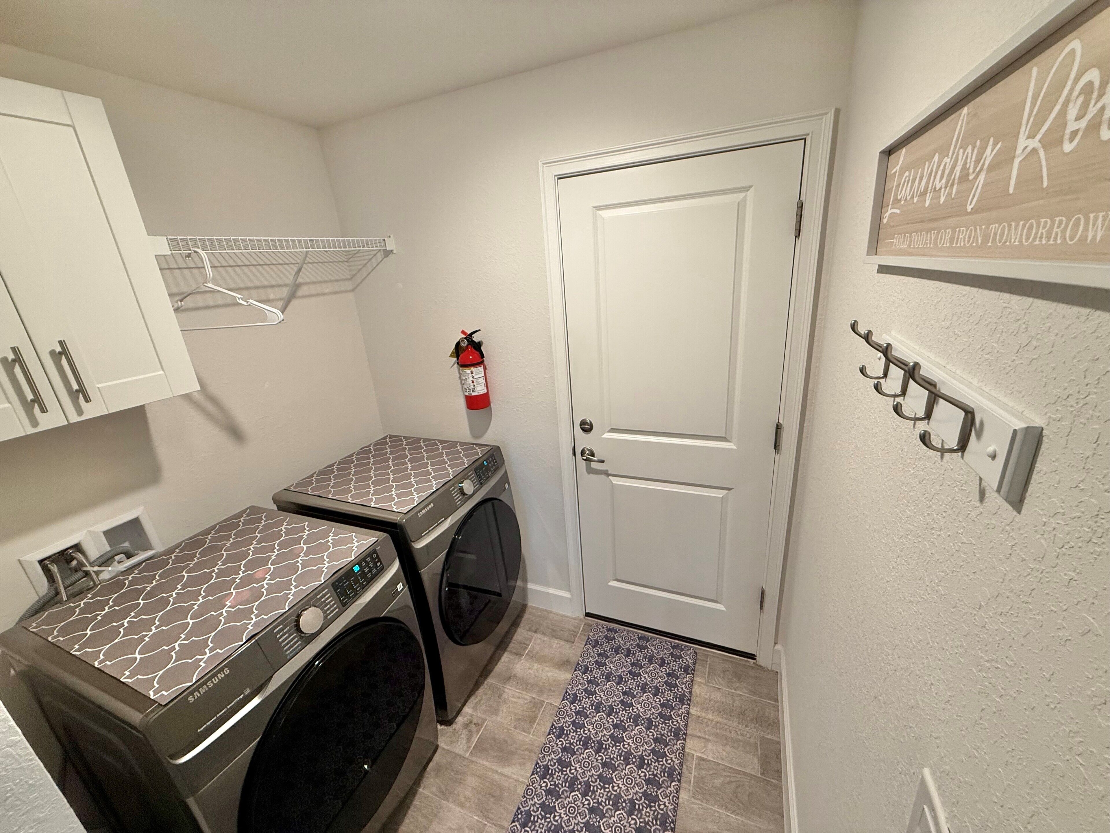 Una sala de lavandería con dos lavadoras Samsung, un extintor, una puerta y un perchero en la pared.
