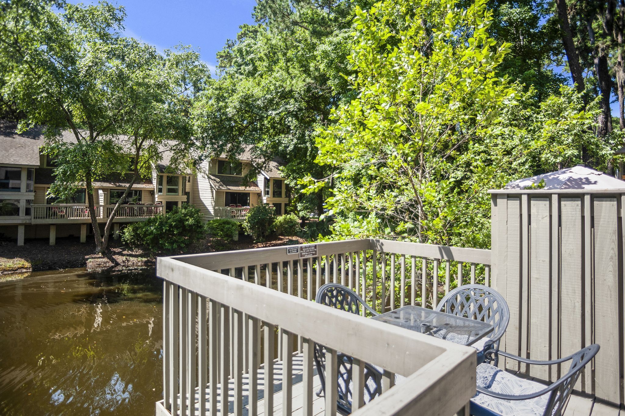 A wooden deck with metal chairs overlooking a pond, surrounded by lush greenery and a building with multiple balconies.