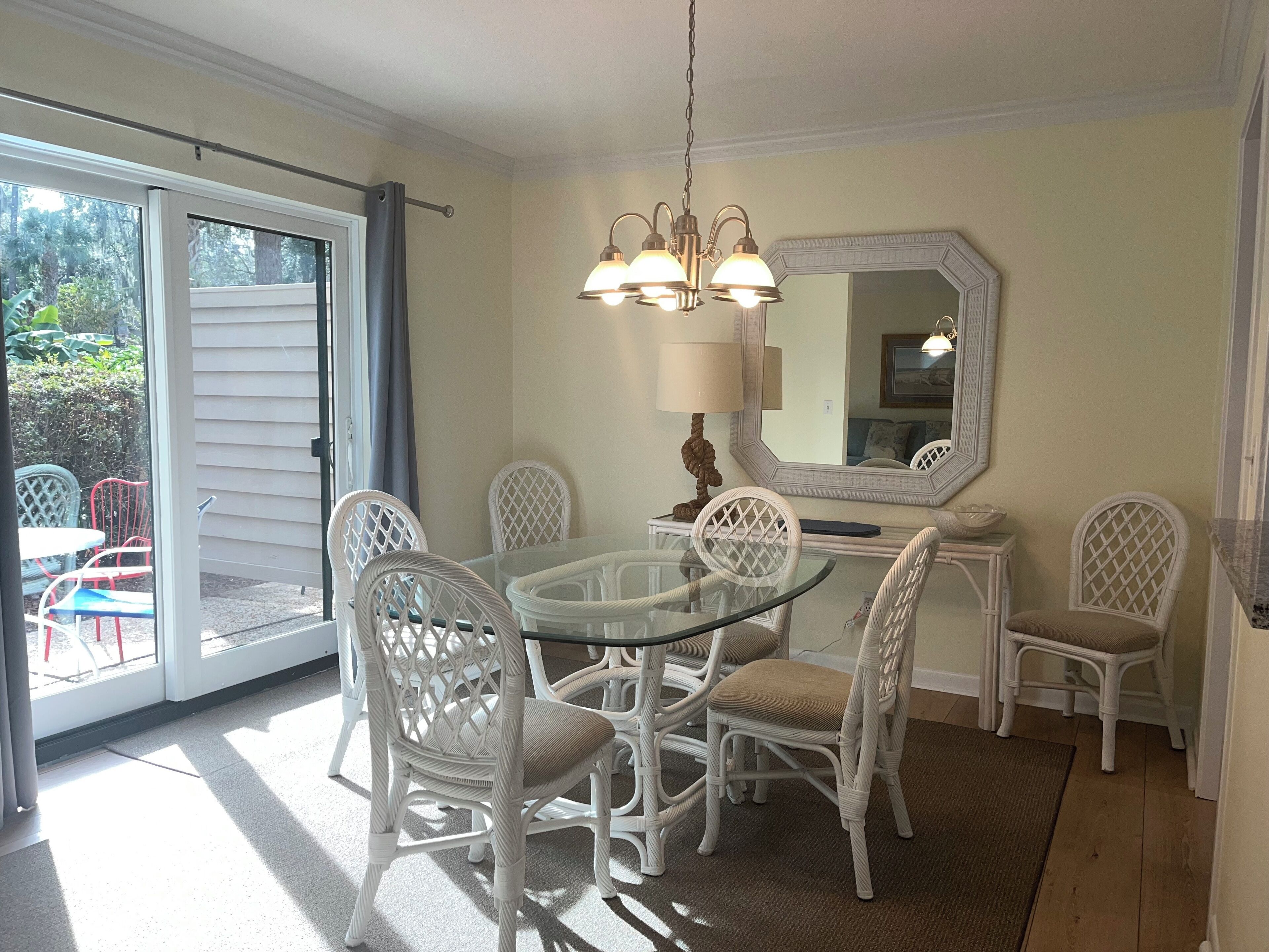 Un coin repas avec une table en verre entourée de chaises en osier blanc, un lustre et un grand miroir au mur.