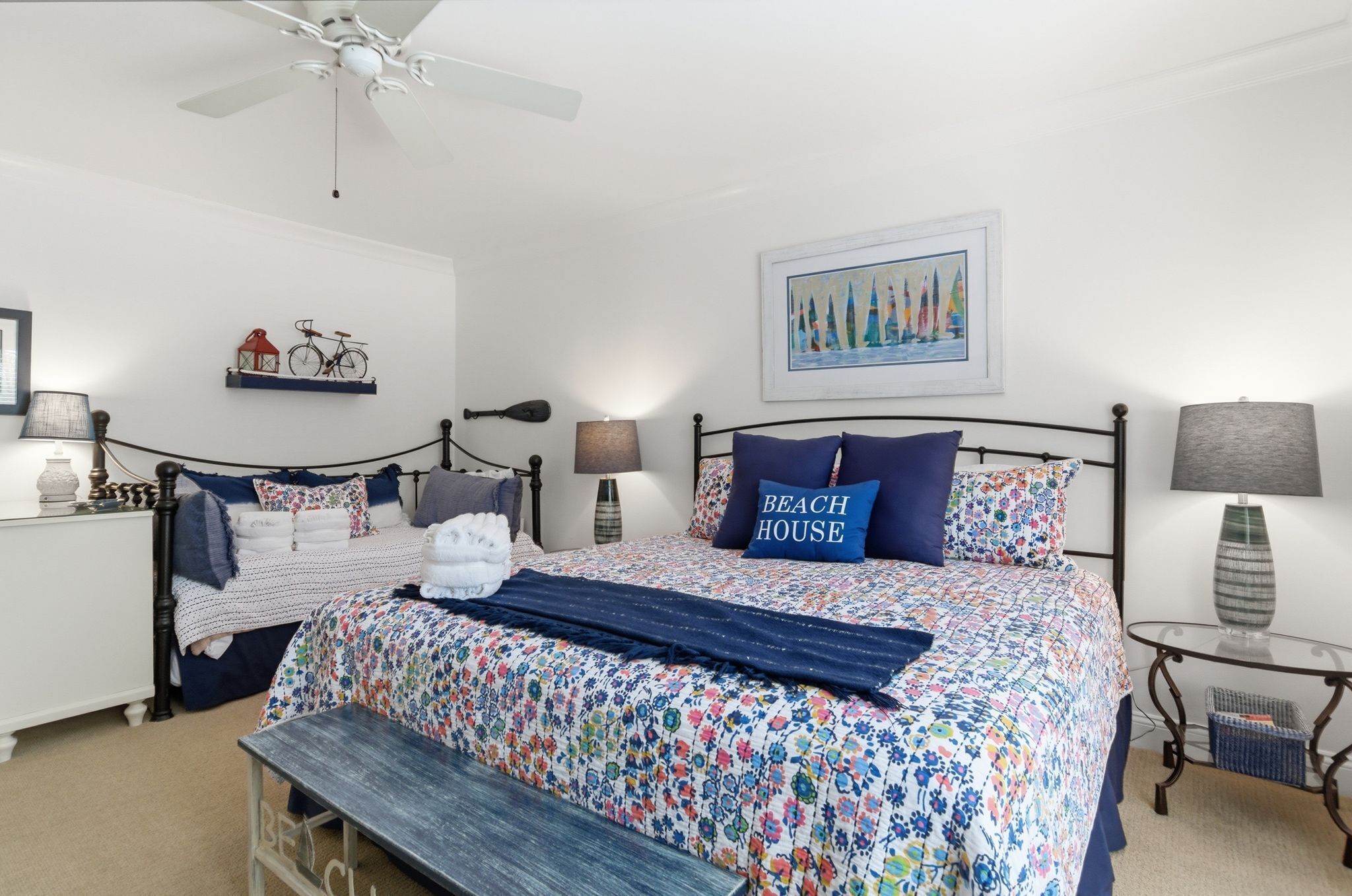 A bedroom with a bed, bedside tables, a bench, a ceiling fan, and a wall-mounted shelf with decorative items.