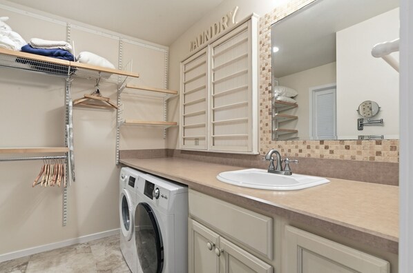 A laundry room with a washing machine, shelves with folded clothes, a sink, and a mirror. - 2410 Lighthouse Tennis at The Sea Pines Resort (Hilton Head Island)