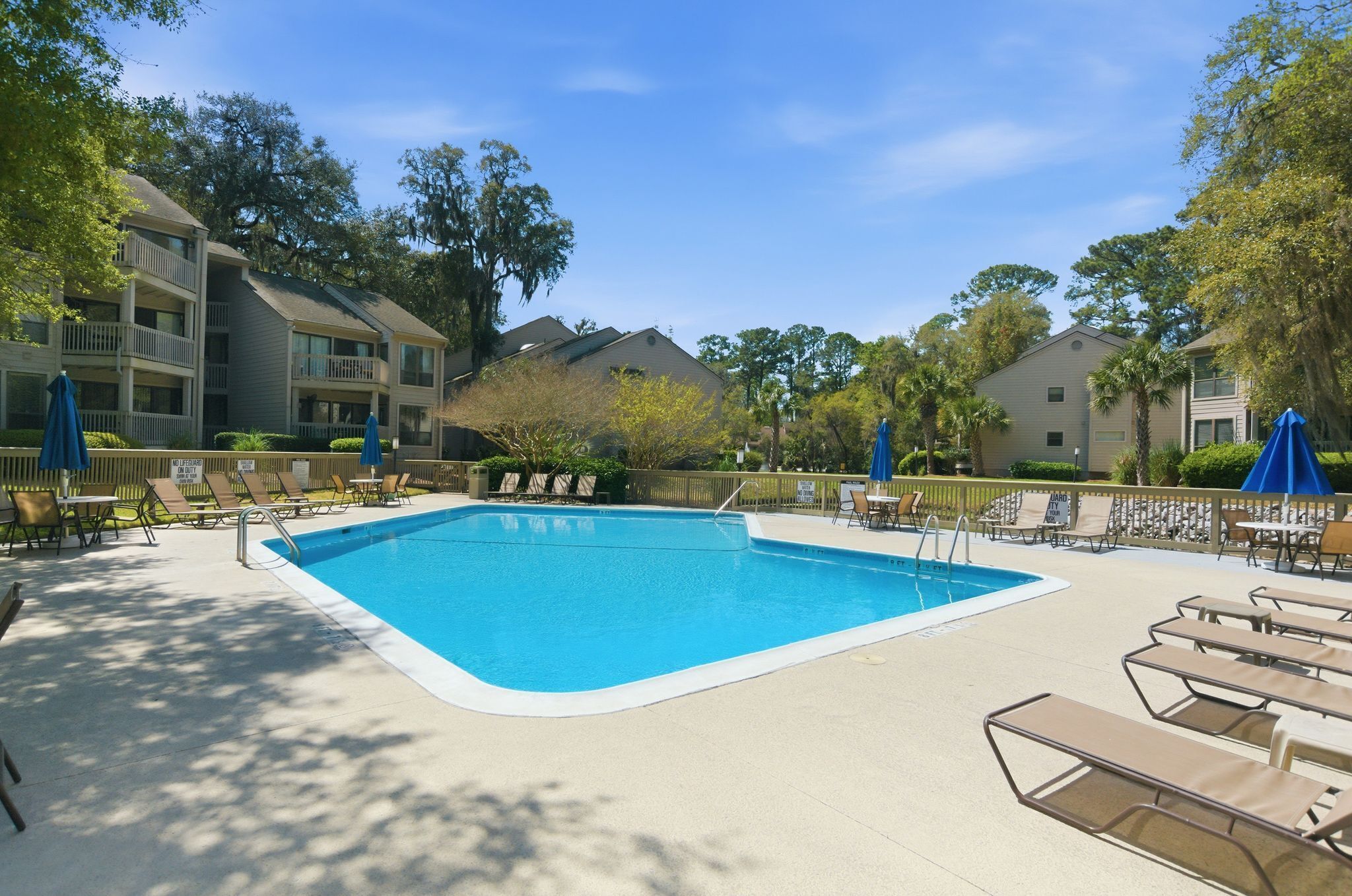 A swimming pool with lounge chairs and umbrellas, surrounded by trees and multi-story buildings.
