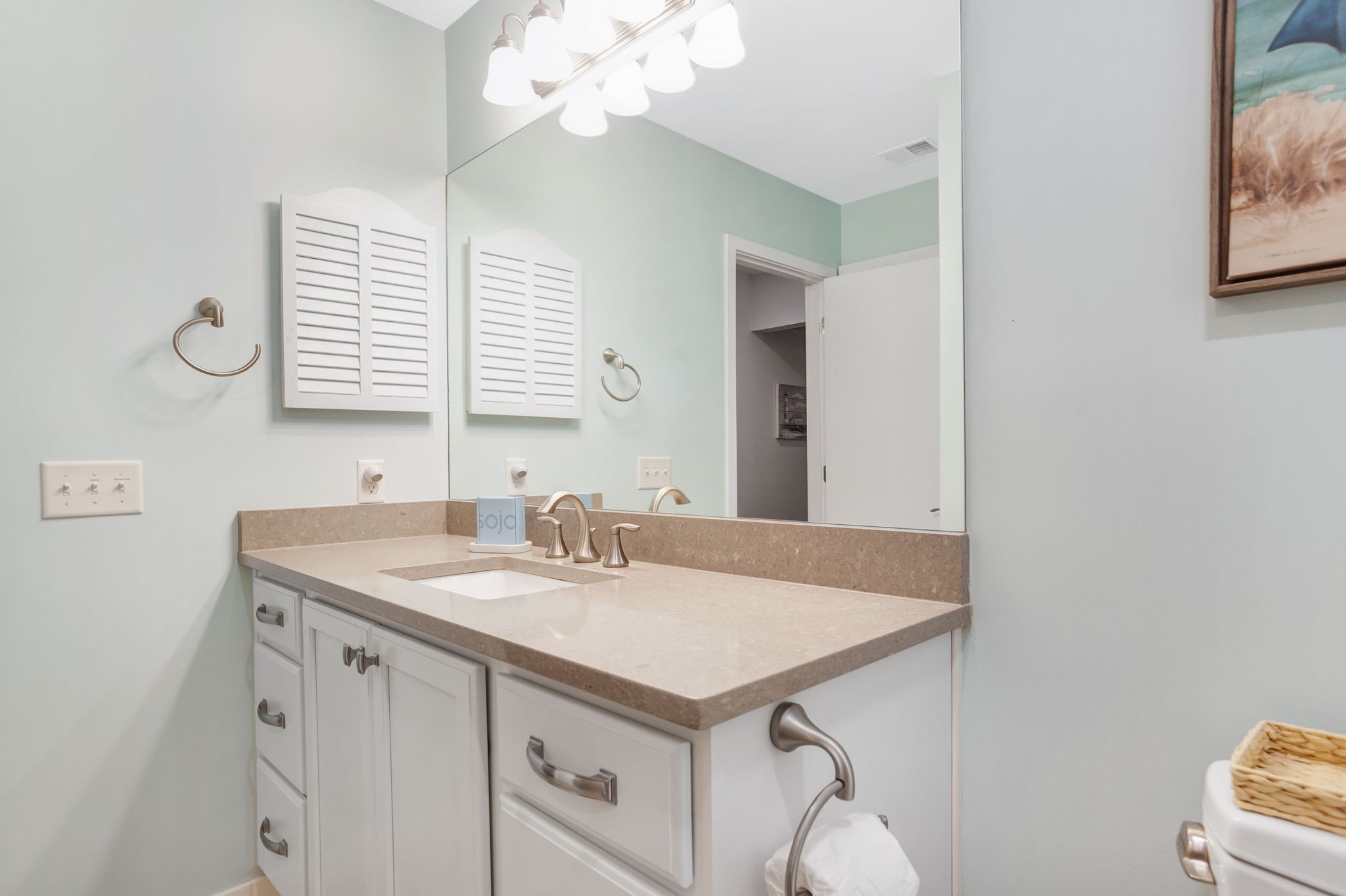 A bathroom with a beige countertop, a sink, a mirror, and a toilet. There are two towel rings on the wall, and a framed picture is visible on the wall.