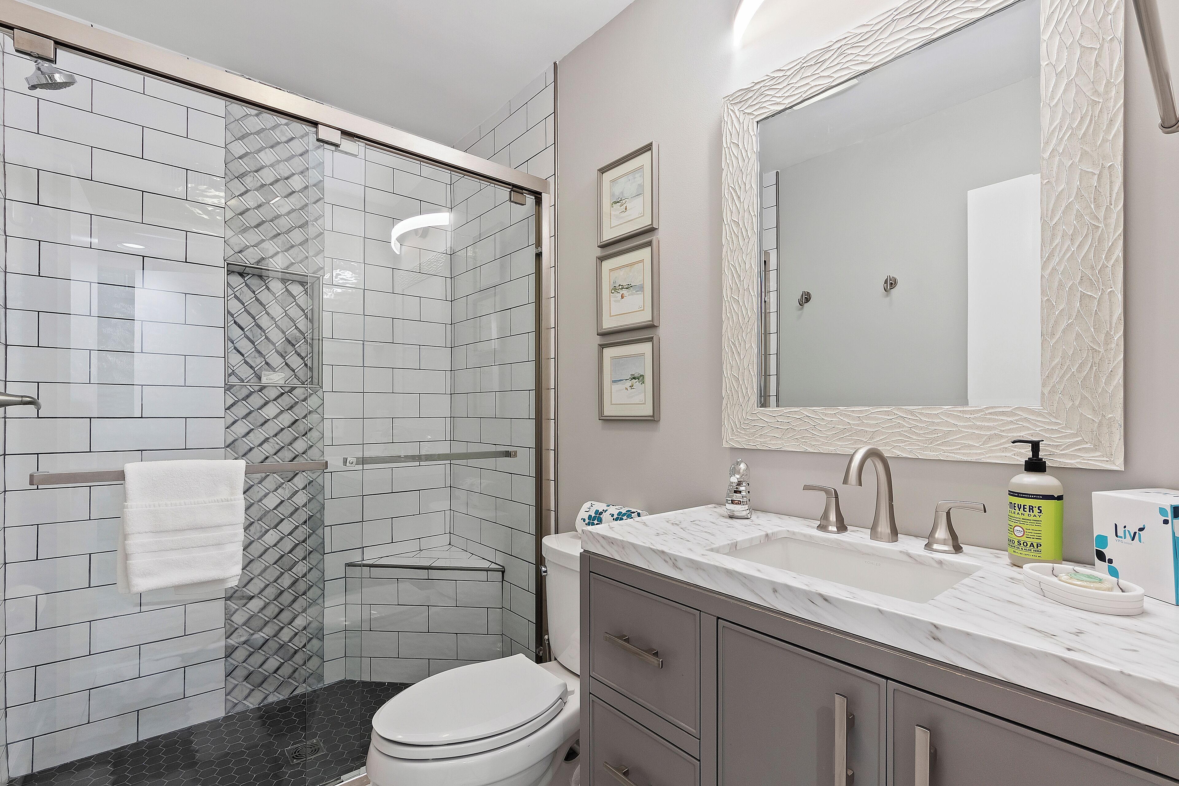 A modern bathroom with a glass shower enclosure, a white toilet, a marble sink with a large mirror, and a vanity with drawers.