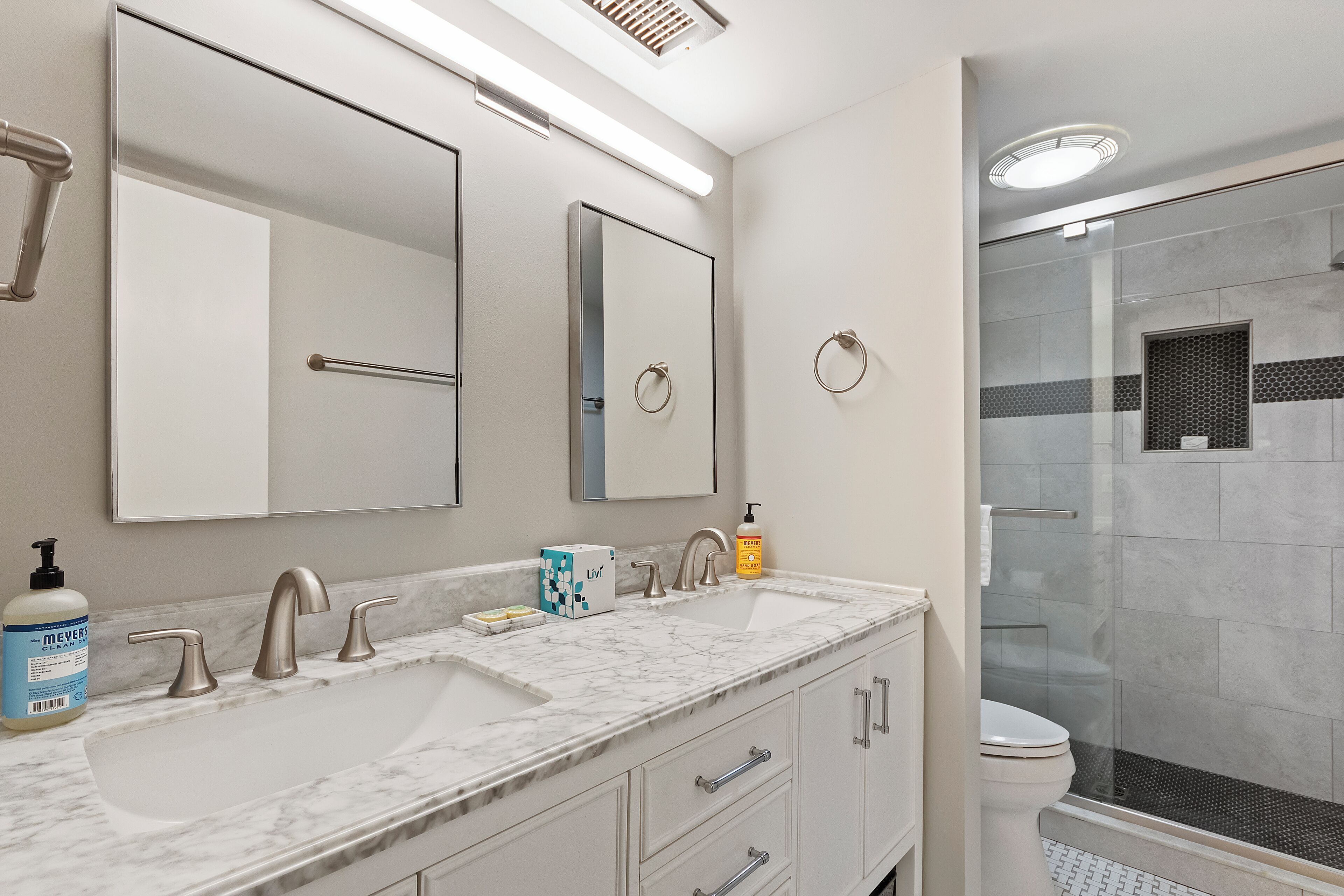 A modern bathroom with a large mirror, two sinks, a toilet, and a glass-enclosed shower.
