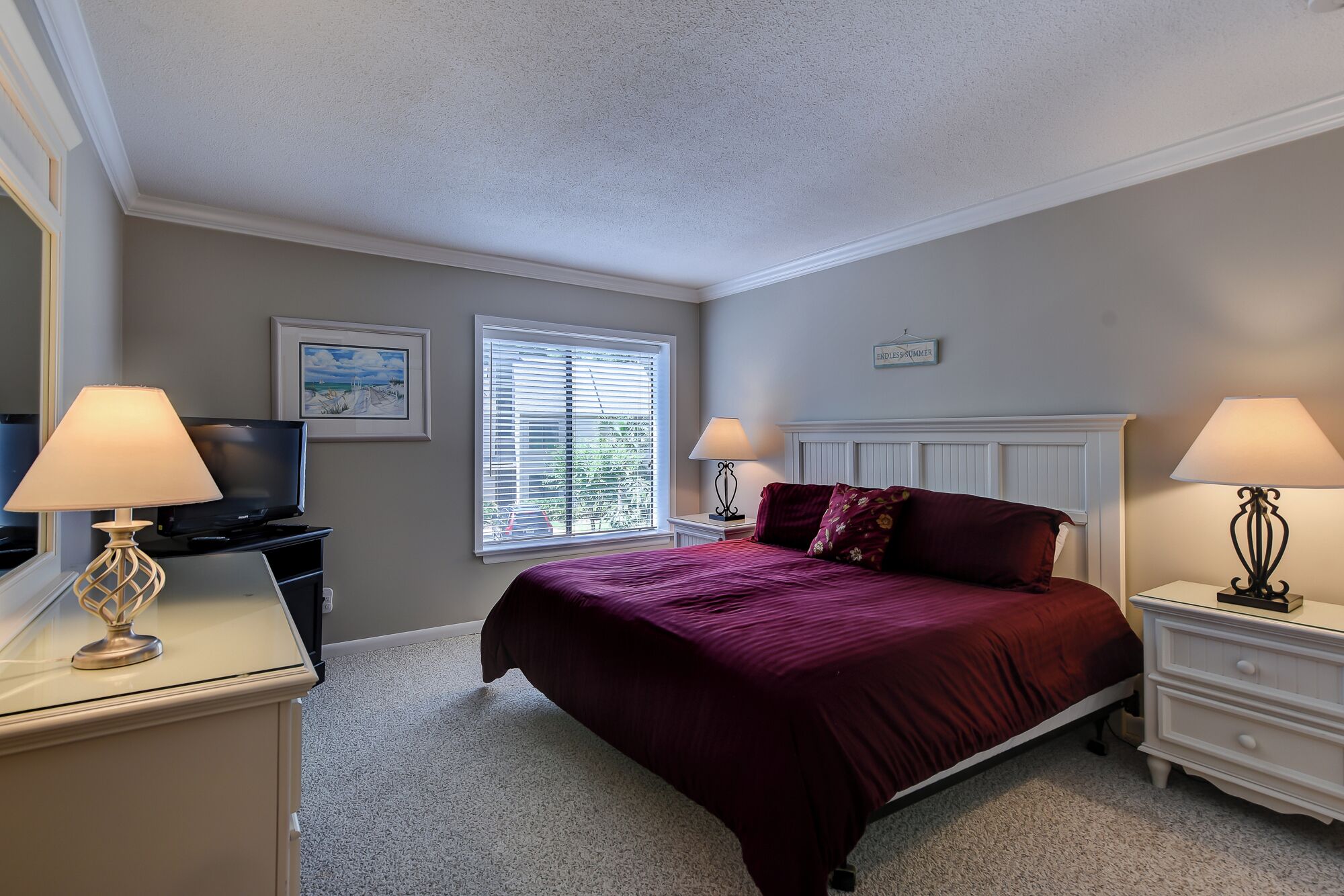 A bedroom with a bed, two bedside tables with lamps, a TV, a window with blinds, and a painting on the wall.