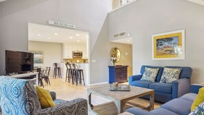 A modern living room with a sofa, armchair, and coffee table. A kitchen is visible in the background with a sign reading "BEACH". - 2302 Heritage Villas at The Sea Pines Resort (Hilton Head Island)