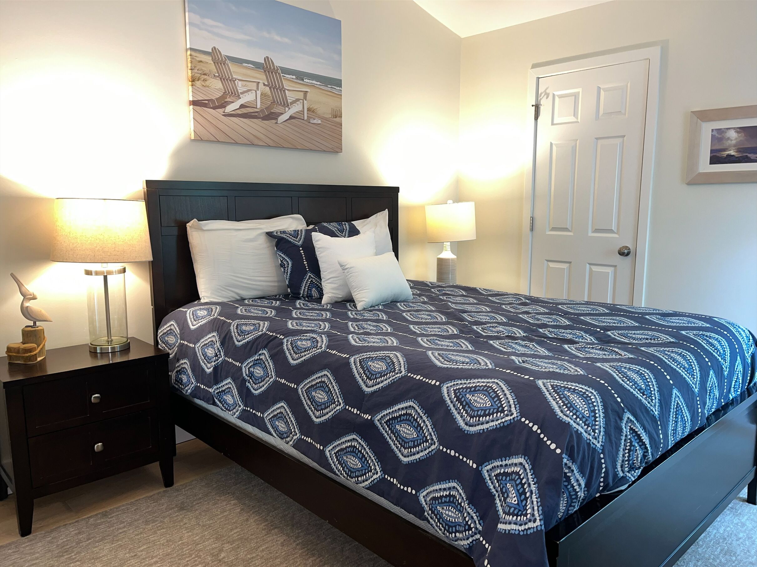 A neatly made bed with a patterned bedspread, two bedside lamps, a nightstand with a decorative item, and a painting above the bed.
