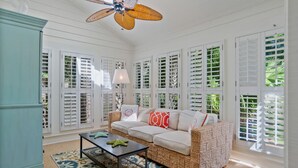 Une pièce avec un canapé blanc, un ventilateur de plafond et de grandes fenêtres équipées de volets.