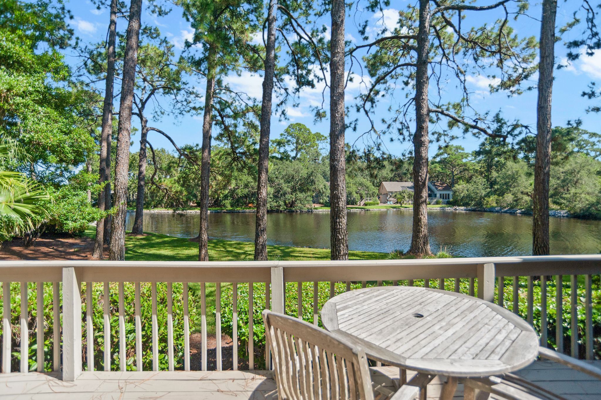 A wooden deck with a table and chairs overlooking a lake with tall trees and a house in the background.