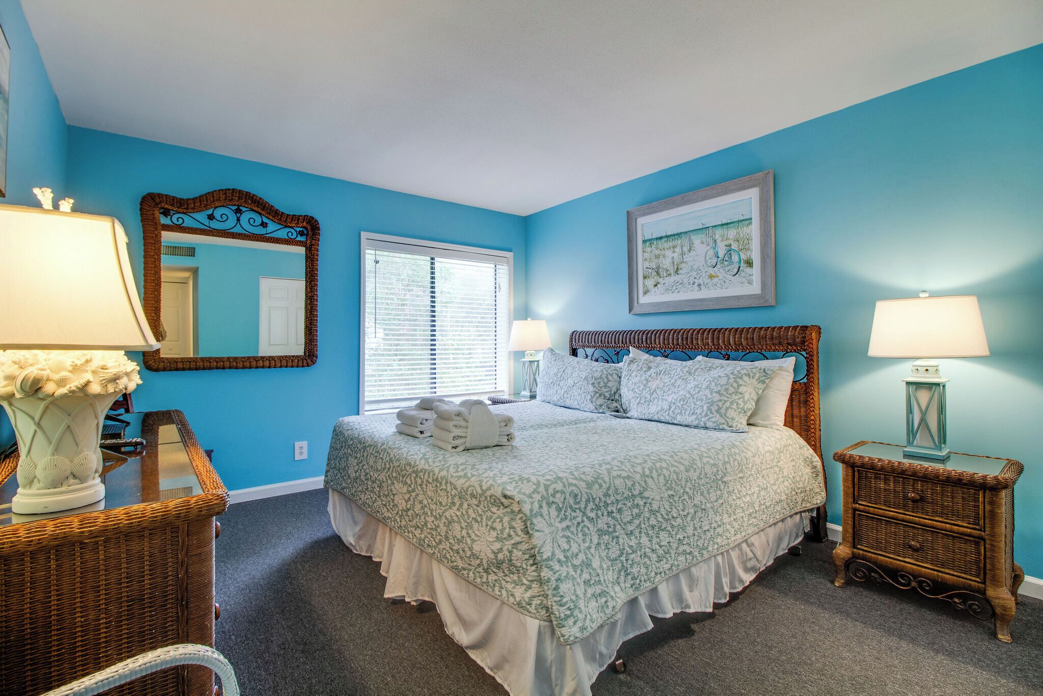 A bedroom with a large bed, two bedside tables with lamps, a mirror, and a window with blinds.