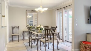 Un comedor con una mesa de vidrio rodeada de sillas ornamentadas, una araña y vistas al exterior a través de amplios ventanales.