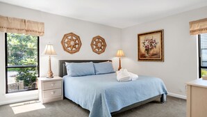 A bedroom with a bed, two bedside lamps, a nightstand, a window with curtains, and a painting on the wall. - 2629 Calibogue Club at The Sea Pines Resort (Hilton Head Island)