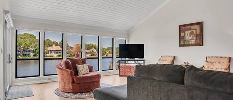 A spacious living room with a large window overlooking a body of water, a flat-screen TV, and a ceiling fan.