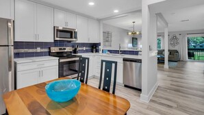 A modern kitchen with white cabinets, a stainless steel refrigerator, a microwave, and a dishwasher. There is a wooden dining table with a blue bowl. - 2350 Racquet Club at The Sea Pines Resort (Hilton Head Island)