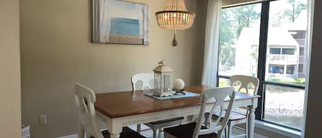 A dining area with a wooden table, white chairs, a blue rug, a chandelier, and a painting of a beach scene.