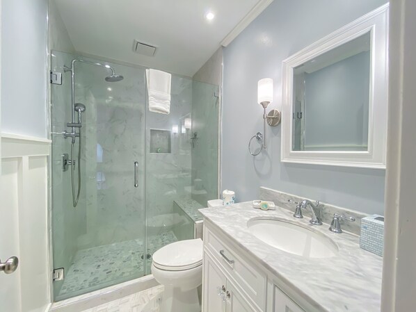 Un baño moderno con ducha de vidrio, inodoro blanco, lavamanos de mármol y un espejo grande.