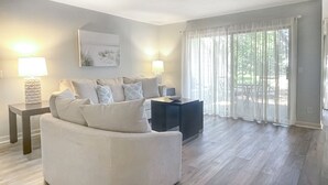 Un salon moderne avec un canapé blanc, deux tables d’appoint avec des lampes, une table basse foncée et une grande fenêtre ornée de rideaux légers.