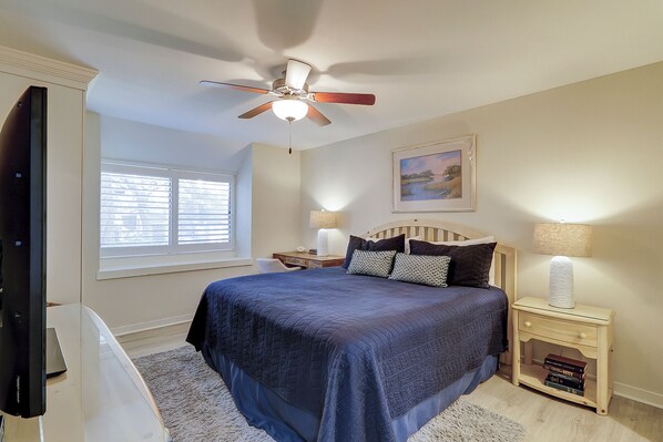 A bedroom with a bed, a ceiling fan, a window with blinds, a bedside table with a lamp, and a framed picture on the wall.
