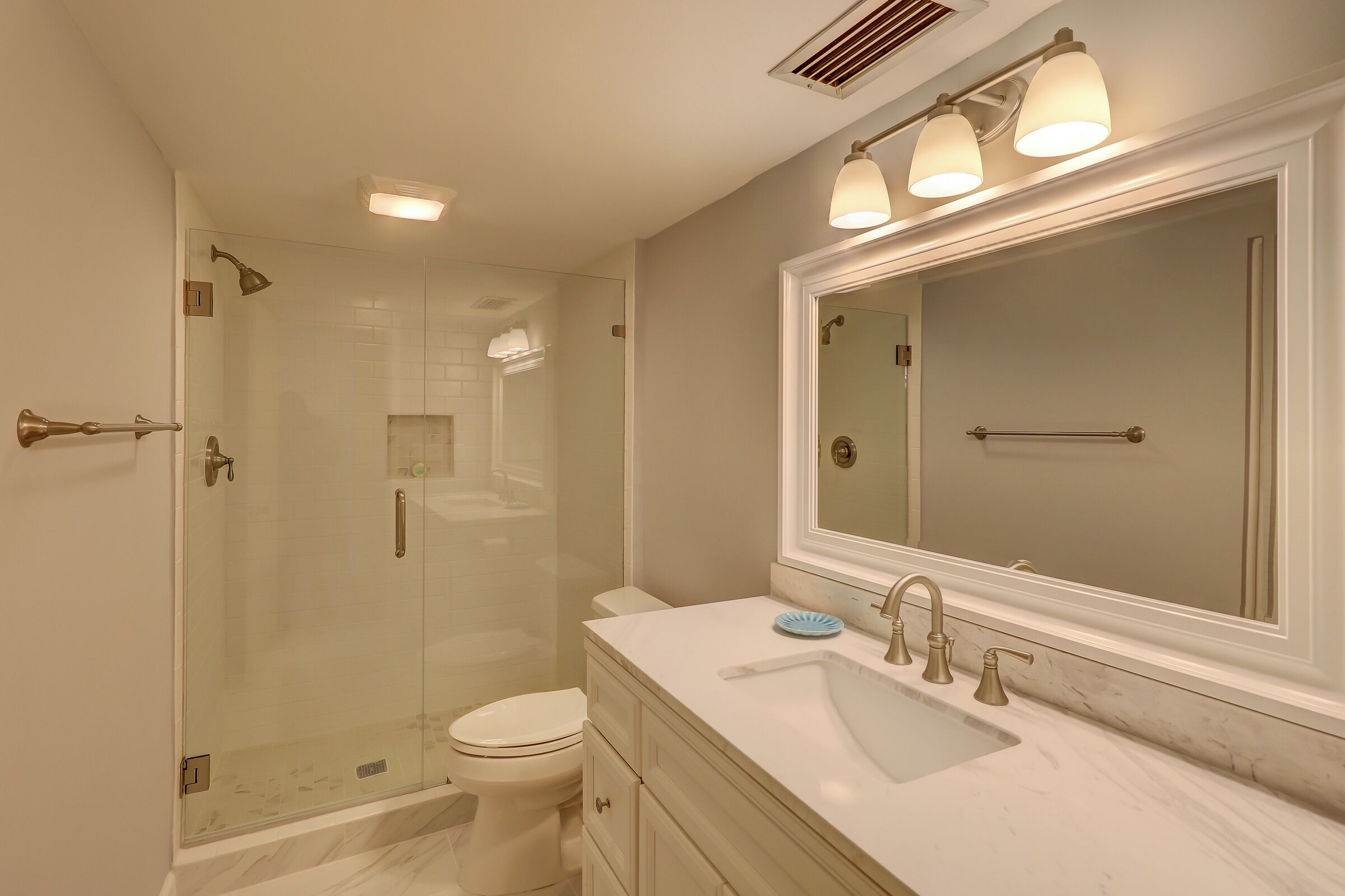 A bathroom with a glass-enclosed shower, a toilet, a sink with a large mirror, and a vanity with drawers.