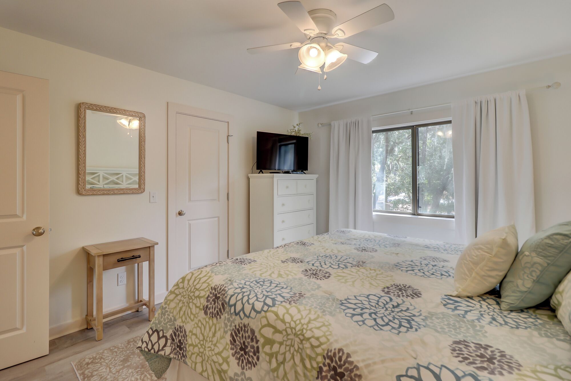 A bedroom with a bed, a TV on a dresser, a ceiling fan, and a window with curtains.
