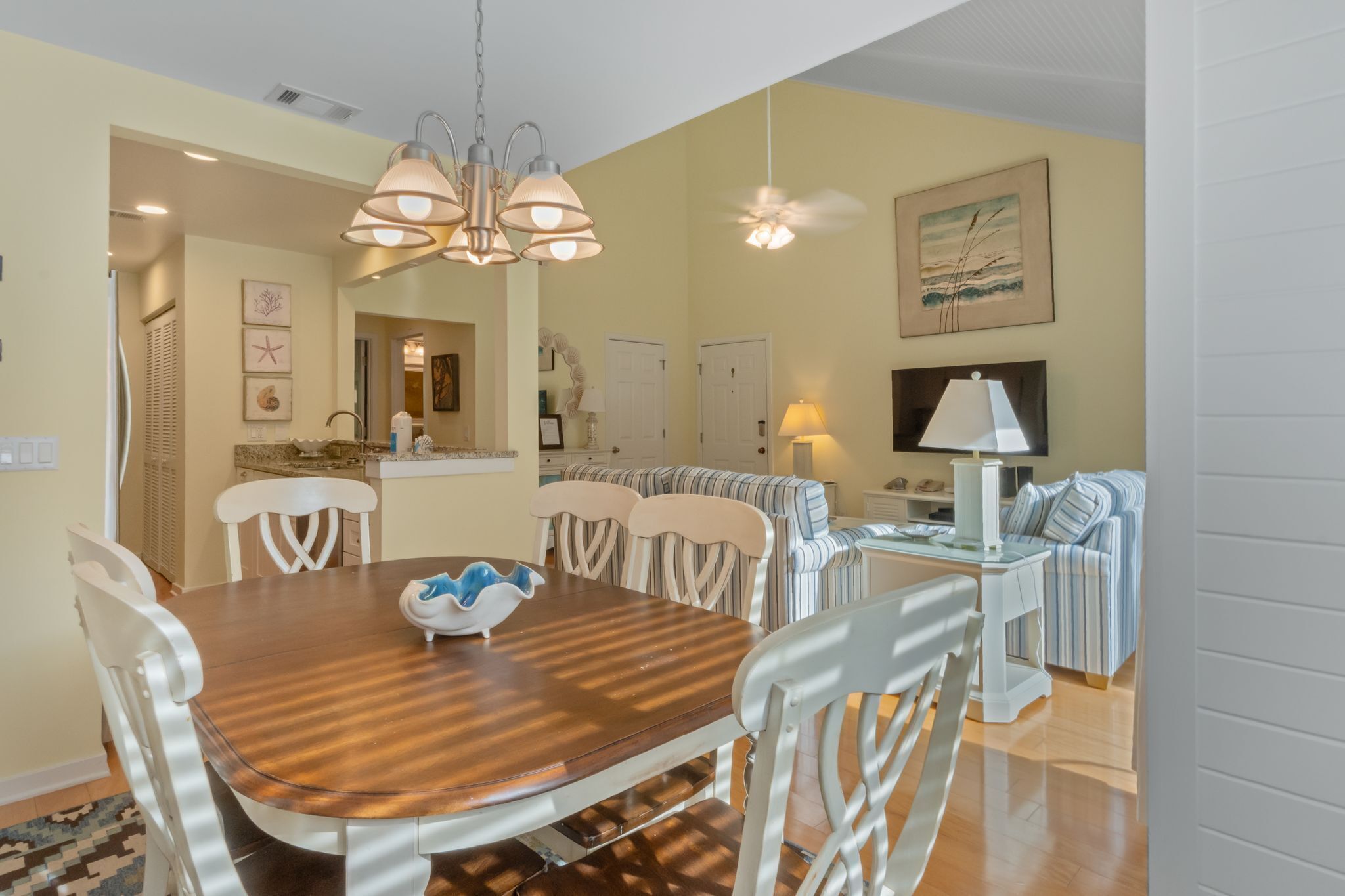 A dining area with a wooden table and white chairs, a ceiling fan, and a kitchen in the background.