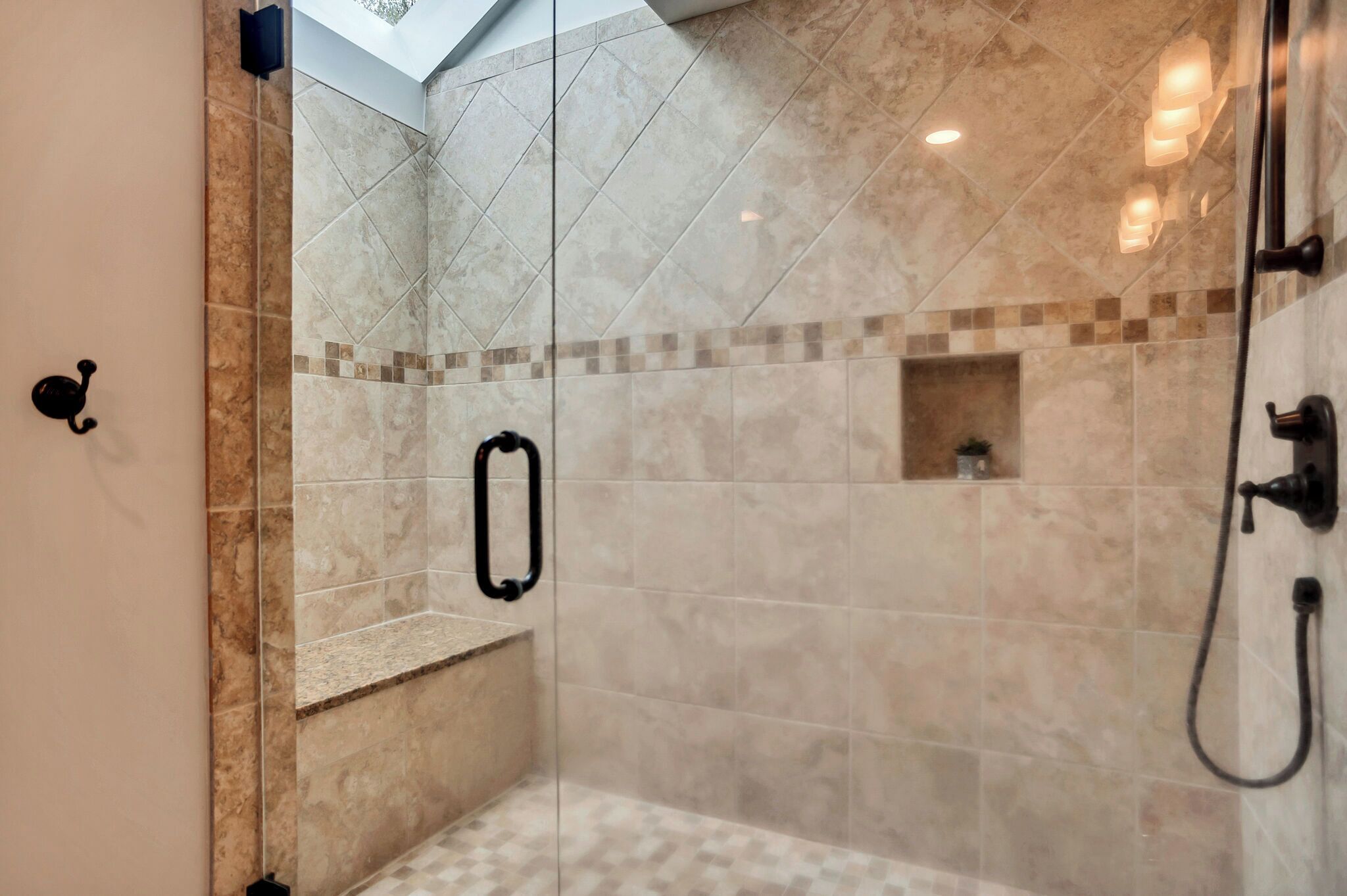 Ducha a pie con puerta de cristal, azulejos color beige, un pequeño banco y un tragaluz en el techo.
