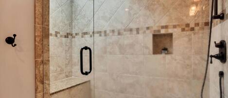 Ducha a pie con puerta de cristal, azulejos color beige, un pequeño banco y un tragaluz en el techo.