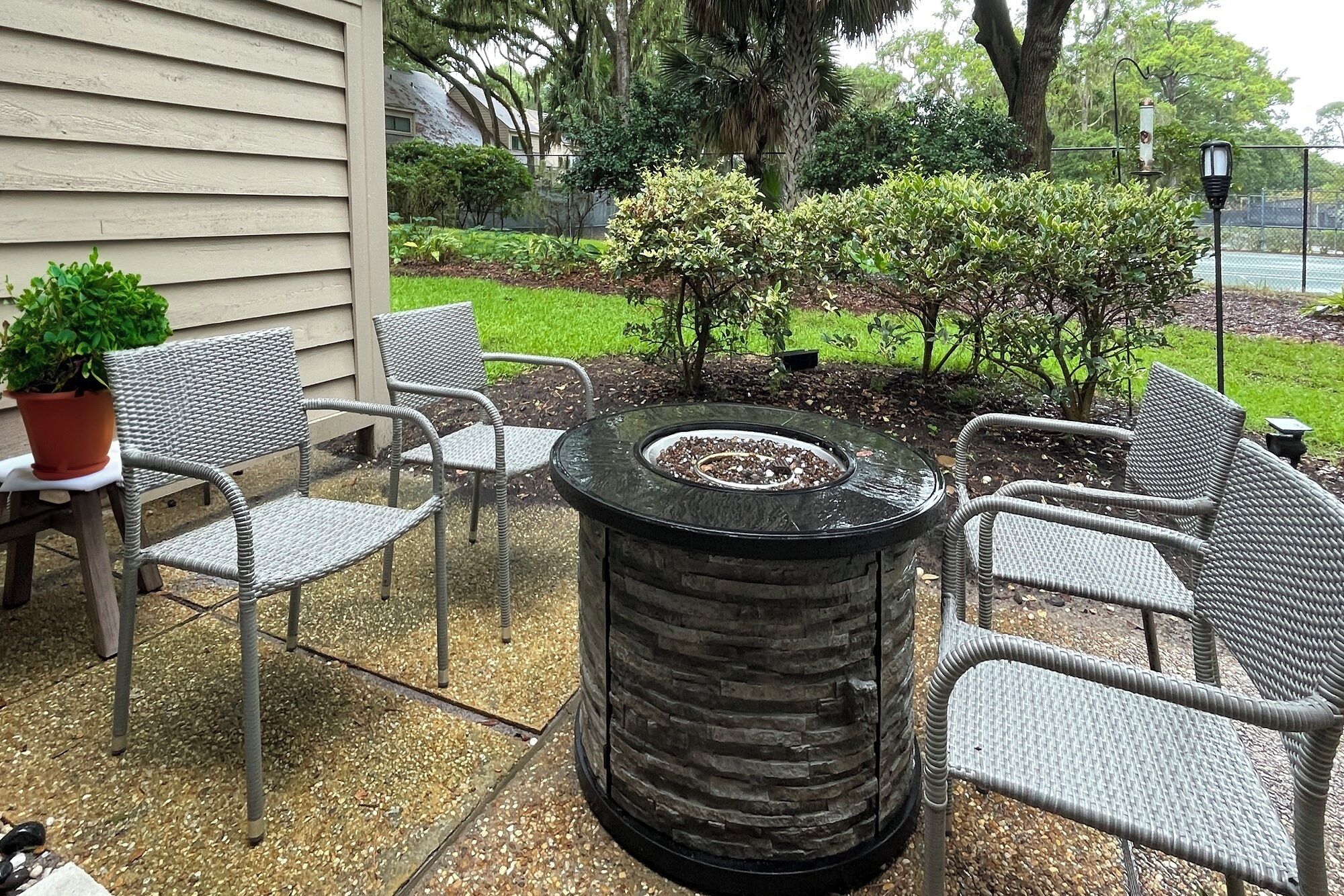 Un patio al aire libre con una mesa de fuego de piedra rodeada de sillas de mimbre, una planta en maceta y un pequeño jardín con arbustos y un árbol.