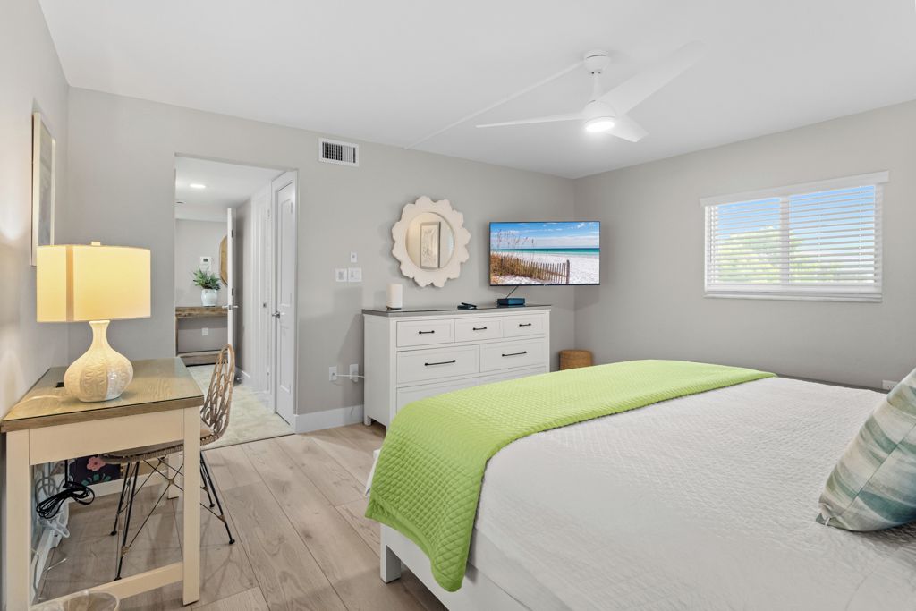 Un dormitorio con cama, escritorio con lámpara, cómoda, televisor y ventana con persianas.