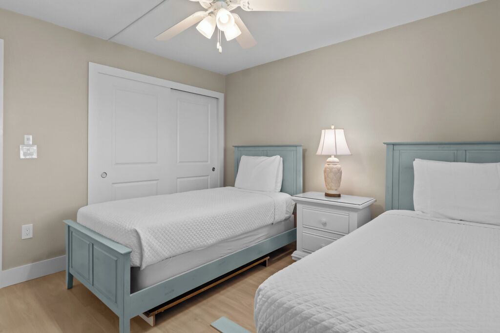 A bedroom with two beds, a ceiling fan, a nightstand with a lamp, and a white door.