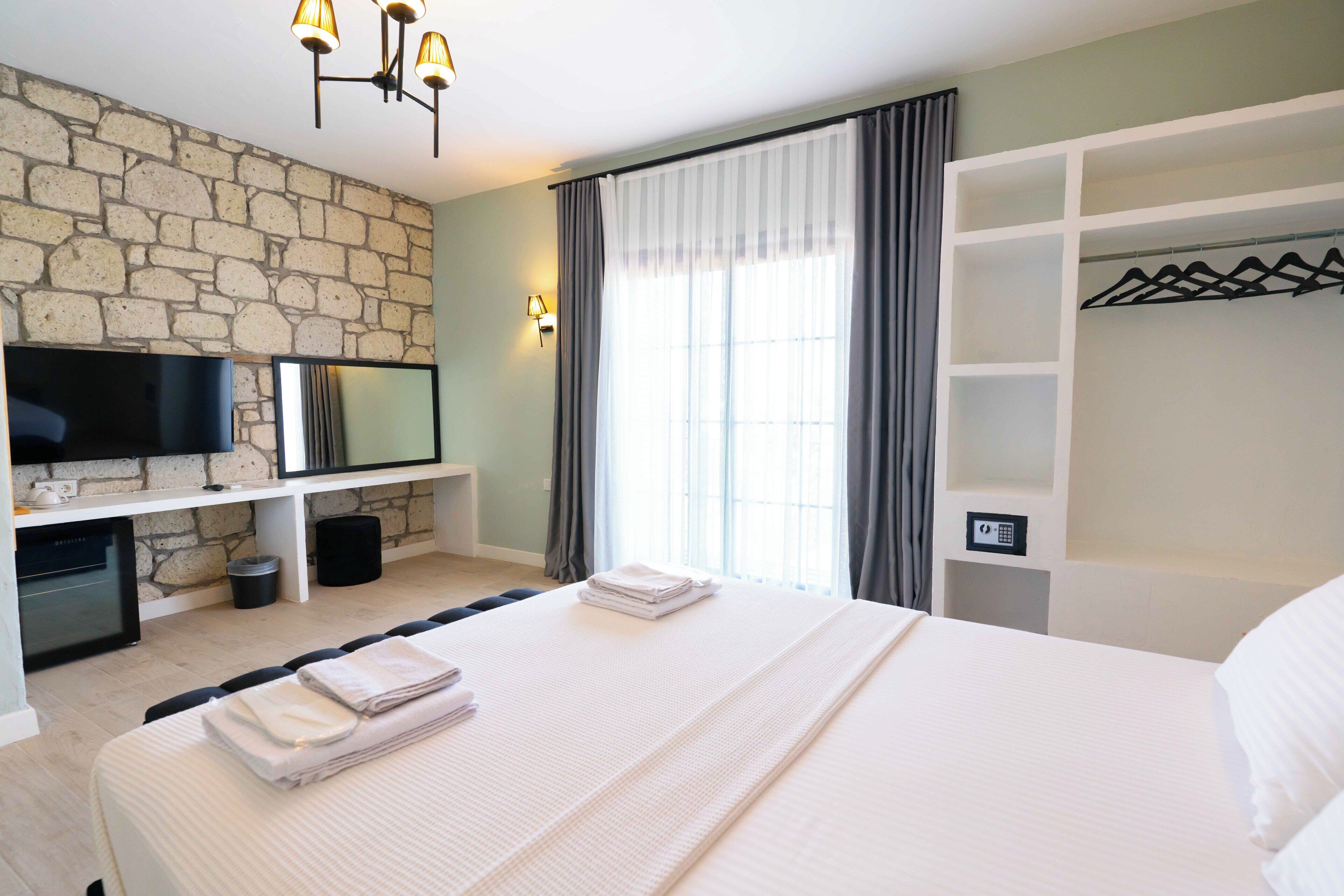 A hotel room with a stone wall, a flat-screen TV, a bed with neatly folded towels, a wardrobe with hangers, and a window with curtains.