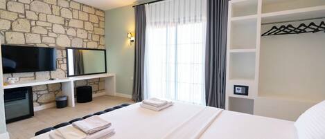 A hotel room with a stone wall, a flat-screen TV, a bed with neatly folded towels, a wardrobe with hangers, and a window with curtains.