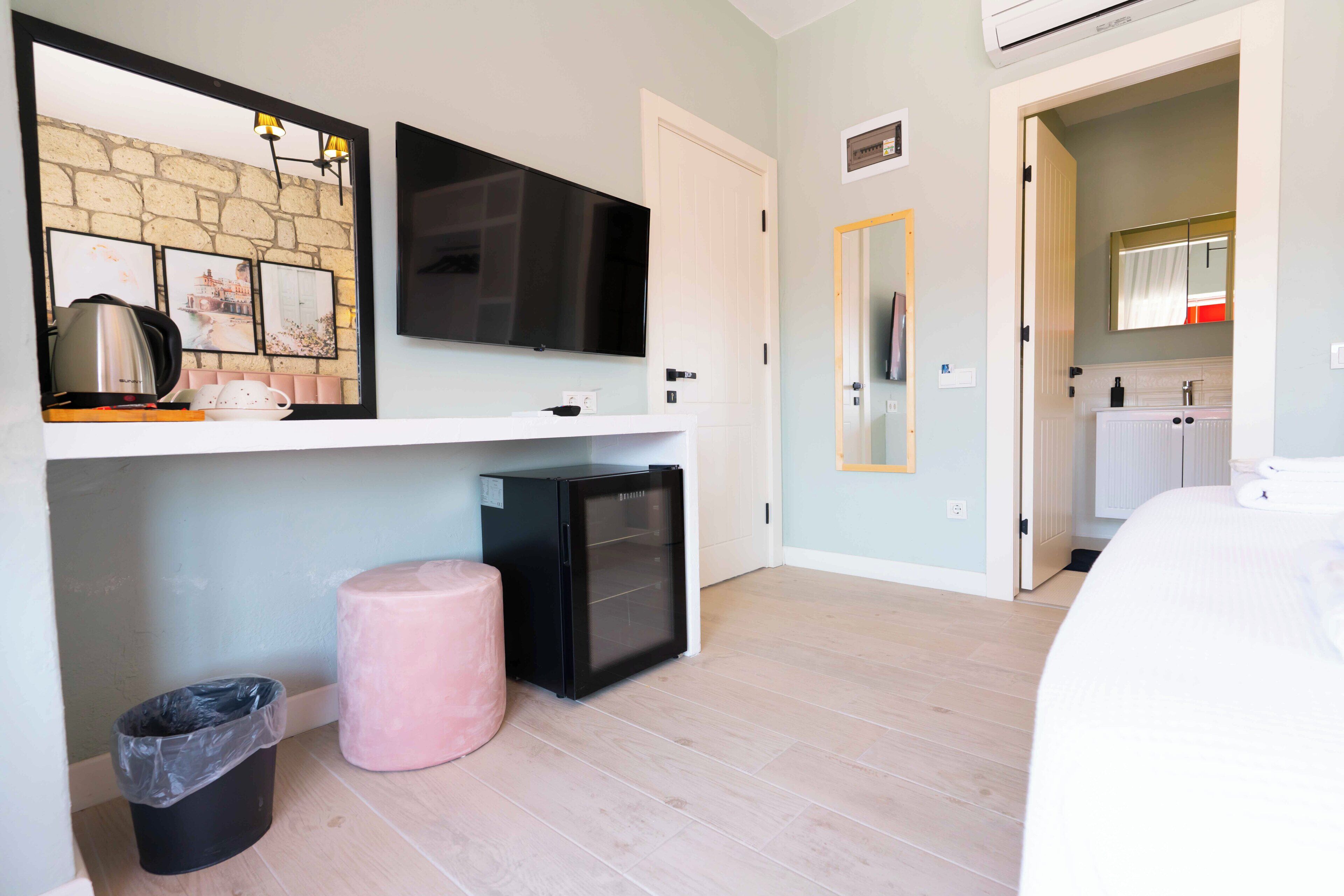 A hotel room with a flat-screen TV mounted above a white console, a small black refrigerator, a pink ottoman, and a trash bin.