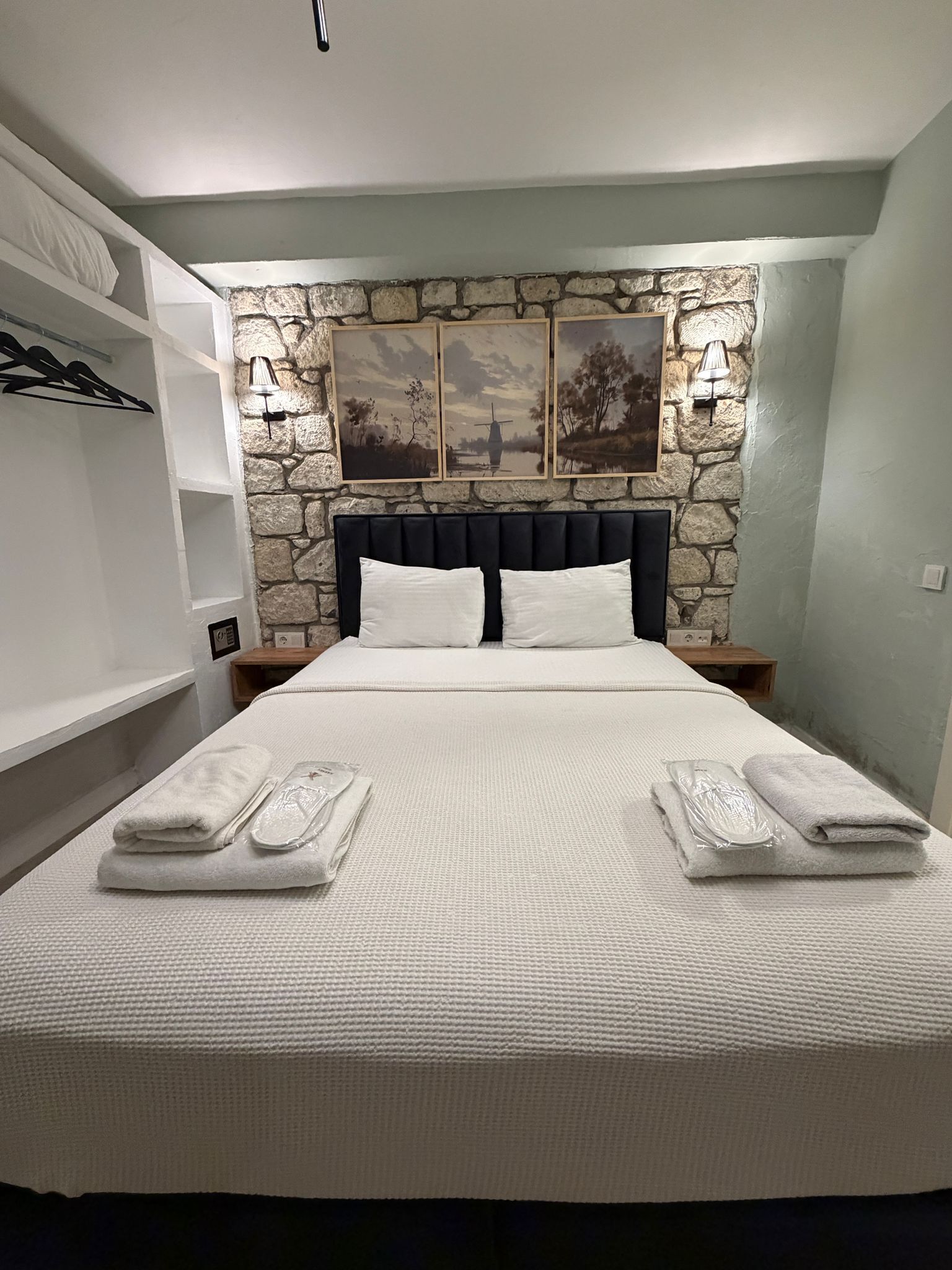 Un lit bien fait, avec du linge de lit blanc et deux oreillers, un mur en pierre orné d’œuvres d’art et une étagère avec des objets suspendus.