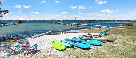 Una playa con kayaks y sillas de descanso, un muelle que se adentra en el agua, y un cielo despejado.